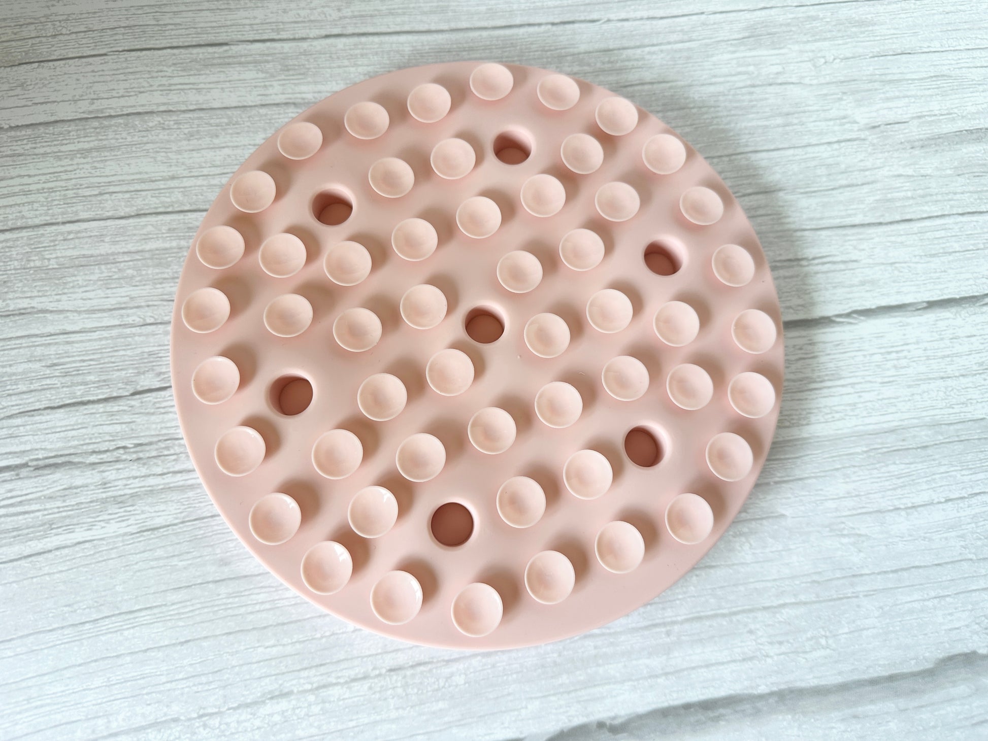 The Crafts by Kate Slow Feeder Pet Mat, a round, pink silicone trivet with raised and cut-out circles and subtle suction cups for stability, rests on a light wood surface.