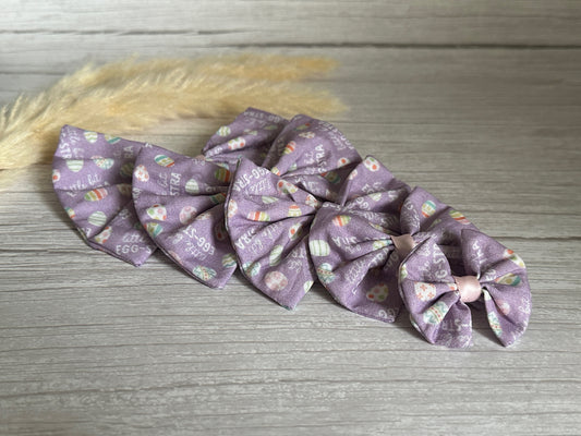 A series of five purple Cotton Pet Bows with collar attachments - Egg-stra from Crafts by Kate, decorated with small Easter-themed designs like eggs, bunnies, and "egg hunt," are arranged in a line from largest to smallest against a wooden background. Made from Egg-stra Print Cotton Fabric and secured with an elastic fastening, a beige feathery decorative piece is placed beside them.