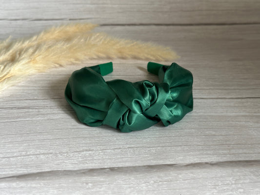 A Silk Knotted Headband from Crafts by Kate in a striking emerald green hue rests gracefully on a wooden surface. To the left, a few soft, beige pampas grass stems add an elegant touch.