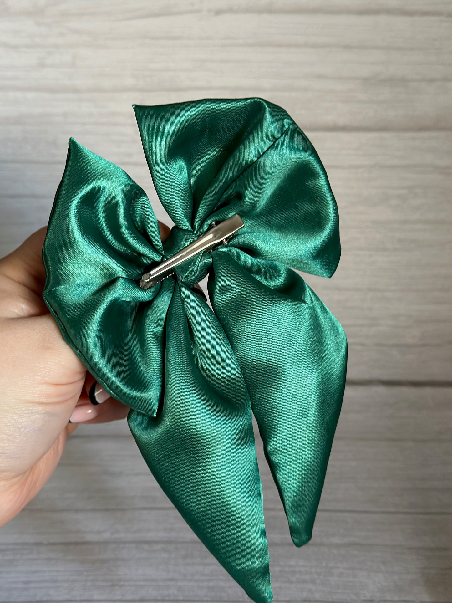 A hand showcases the opulent Silk Sailor Bow Clip in emerald green by Crafts by Kate, featuring a handcrafted satin bow with an exquisite silver hair clip on the reverse. This luxury piece shines brilliantly against a light wooden background.