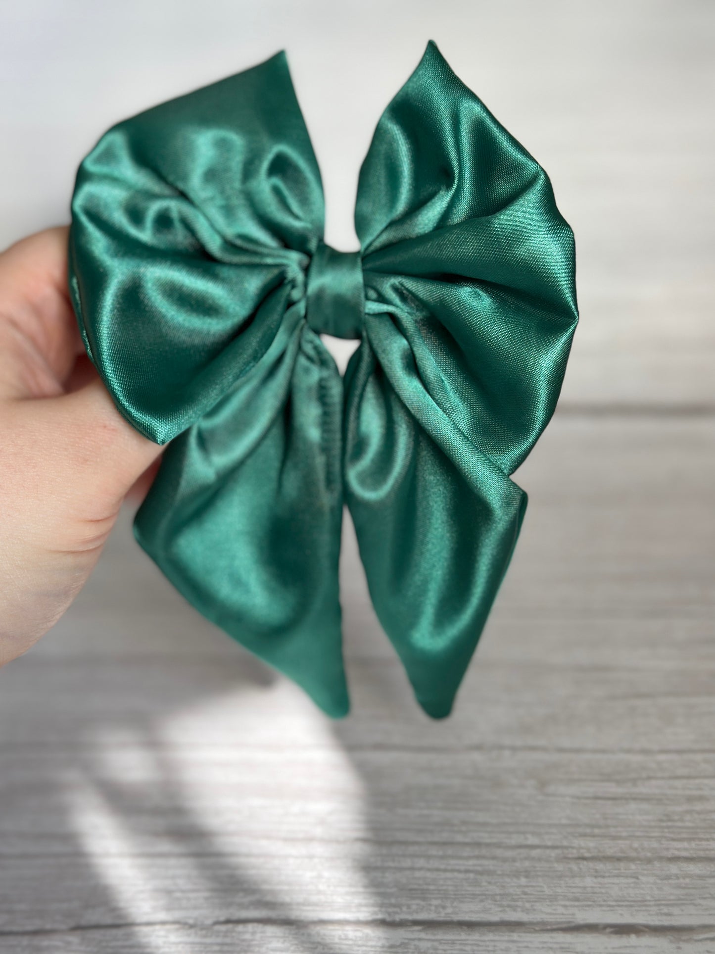 A hand holding the Silk Sailor Bow Clip in emerald green by Crafts by Kate, gleaming against a light gray wooden background.