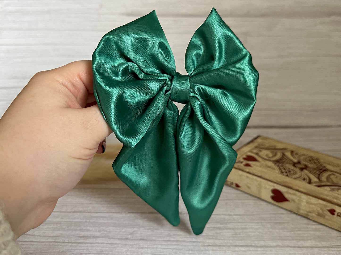 A hand holds the exquisite Silk Sailor Bow Clip in a lush Emerald Green from Crafts by Kate against a light wood background. A handcrafted wooden box embellished with heart designs is partially visible on the side.