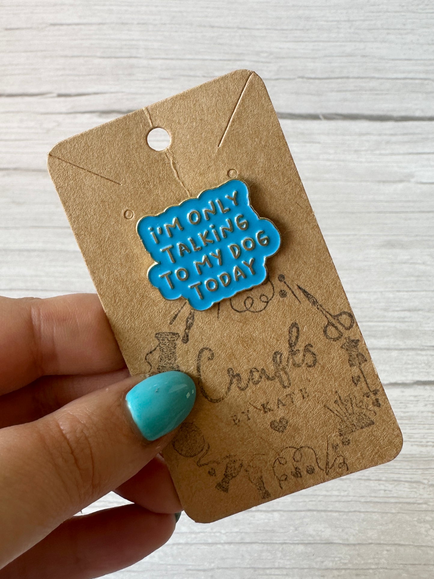A hand with blue-painted nails holds a brown card displaying the "Strawberries & Daisies - Arnie & Mac’s Goodies" enamel pin by Crafts by Kate, set against a light wood background—perfect for dog lovers and peanut butter treat fans.