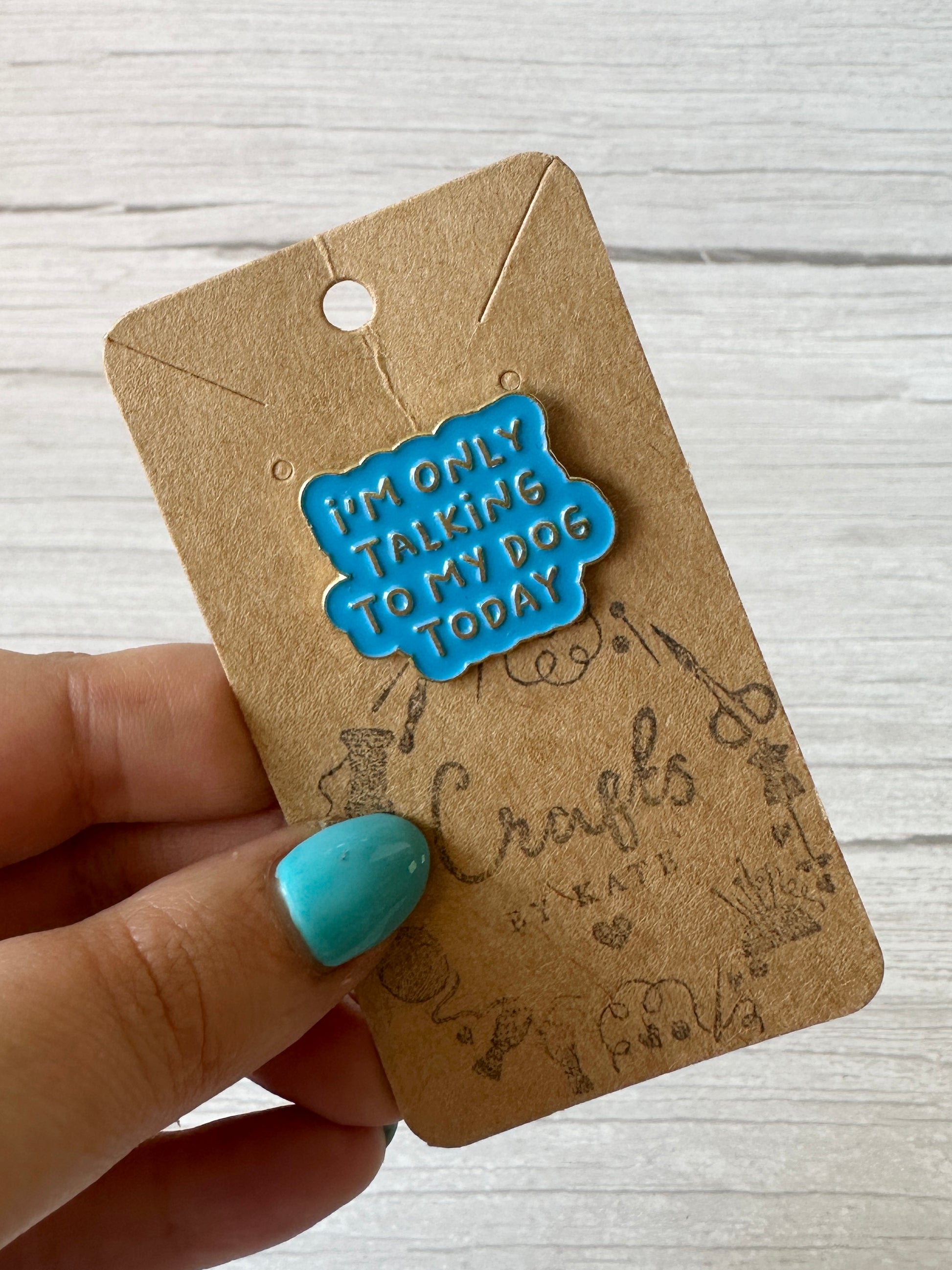 A hand with blue-painted nails holds a brown card displaying the "Strawberries & Daisies - Arnie & Mac’s Goodies" enamel pin by Crafts by Kate, set against a light wood background—perfect for dog lovers and peanut butter treat fans.