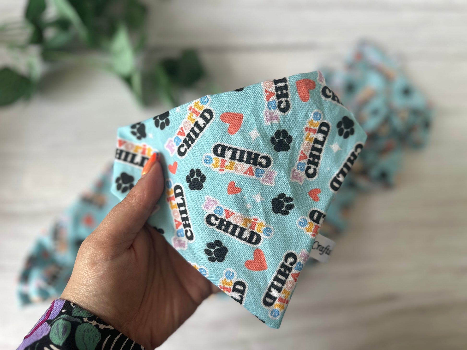 A hand holds the Crafts by Kate Cotton Pet Scrunchie Bandana - Favourite Child, a blue handmade bandana with paw prints, hearts, and playful lettering. A second blurred bandana and a green plant sit in the background on a light surface.