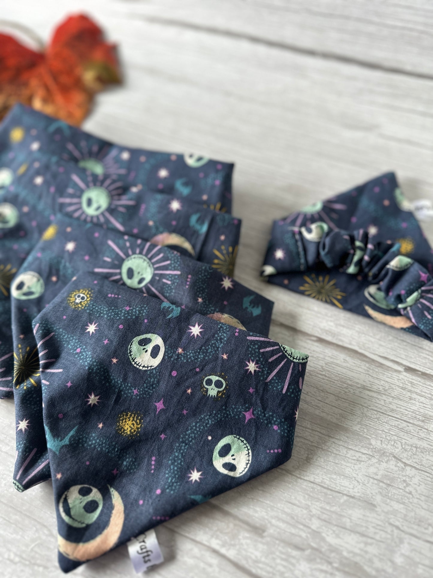 This is a set of dark-themed fabric items adorned with spooky skulls, stars, and moon designs. The set includes carefully folded cloth pieces and a handcrafted "Cotton Pet Scrunchie Bandana - Jack Skeleton" by Crafts by Kate. The items are displayed on a light wooden surface with an orange and black decoration visible in the top left corner.