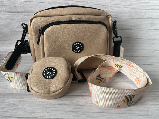 The Bumble Dog Walking Bag by Crafts by Kate is a beige vegan leather crossbody with a matching coin pouch and a cream strap featuring bees and orange flowers, displayed on a light wooden surface.
