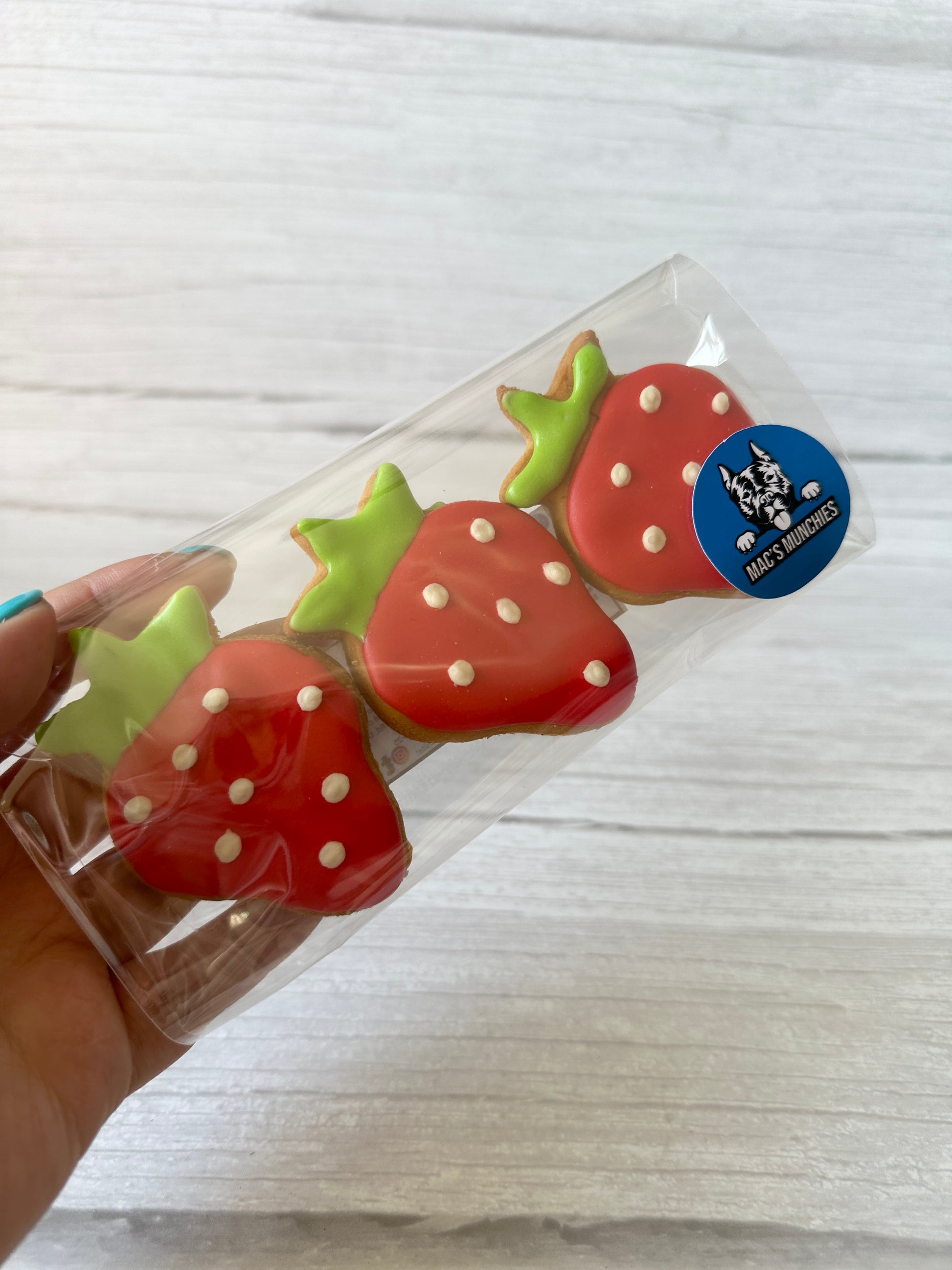 A hand holds a clear box with three strawberry-themed cookies from Crafts by Kate’s Strawberries & Daisies – Arnie & Mac’s Goodies, featuring red icing, white dots, green tops, and a blue sticker—ideal for gifting with pet bandanas or peanut butter treats.