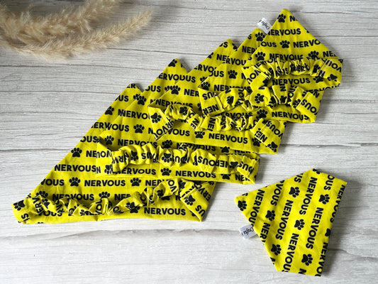 Five "Cotton Pet Scrunchie Bandana - Nervous" by Crafts by Kate, yellow with black paw prints and "NERVOUS" text, are displayed on a light wooden surface with beige pampas grass in the corner.