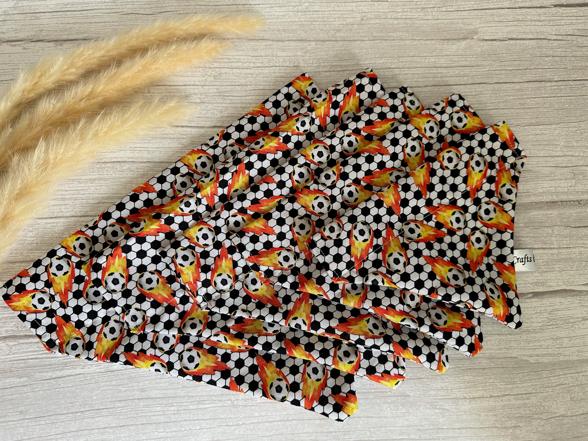 Colorful napkins with a pattern of soccer balls and flames are neatly folded and displayed on a light wood surface. A handcrafted "Cotton Pet Bandana with collar attachment - Football Fan" from Crafts by Kate adds flair, while feathery pampas grass lies to the left of the napkins.