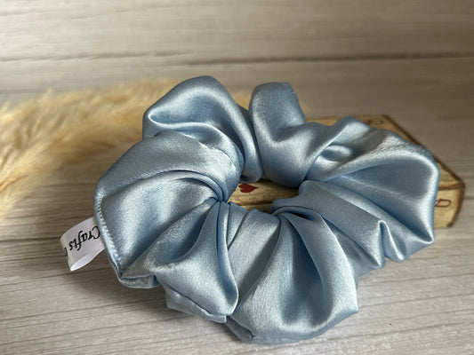 The Silk Extra Large Scrunchie in French Blue by Crafts by Kate sits on a wooden surface next to a beige fluffy object, with a softly blurred rustic wooden box featuring a subtle design in the background.