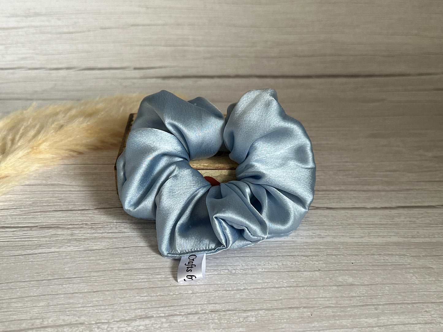 A luxurious Silk Extra Large Scrunchie in French Blue from Crafts by Kate rests elegantly on a wooden surface. Attached is a small tag with text, while a soft, beige feather adds a delicate touch in the background.