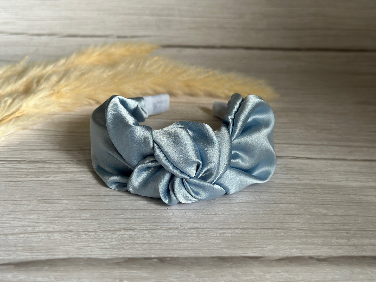 The Silk Knotted Headband - French Blue from Crafts by Kate graces a light wooden surface with its sophisticated twisted design. Carefully handcrafted to perfection, it exudes luxury, enhanced by the natural elegance of soft beige pampas grass in the background.