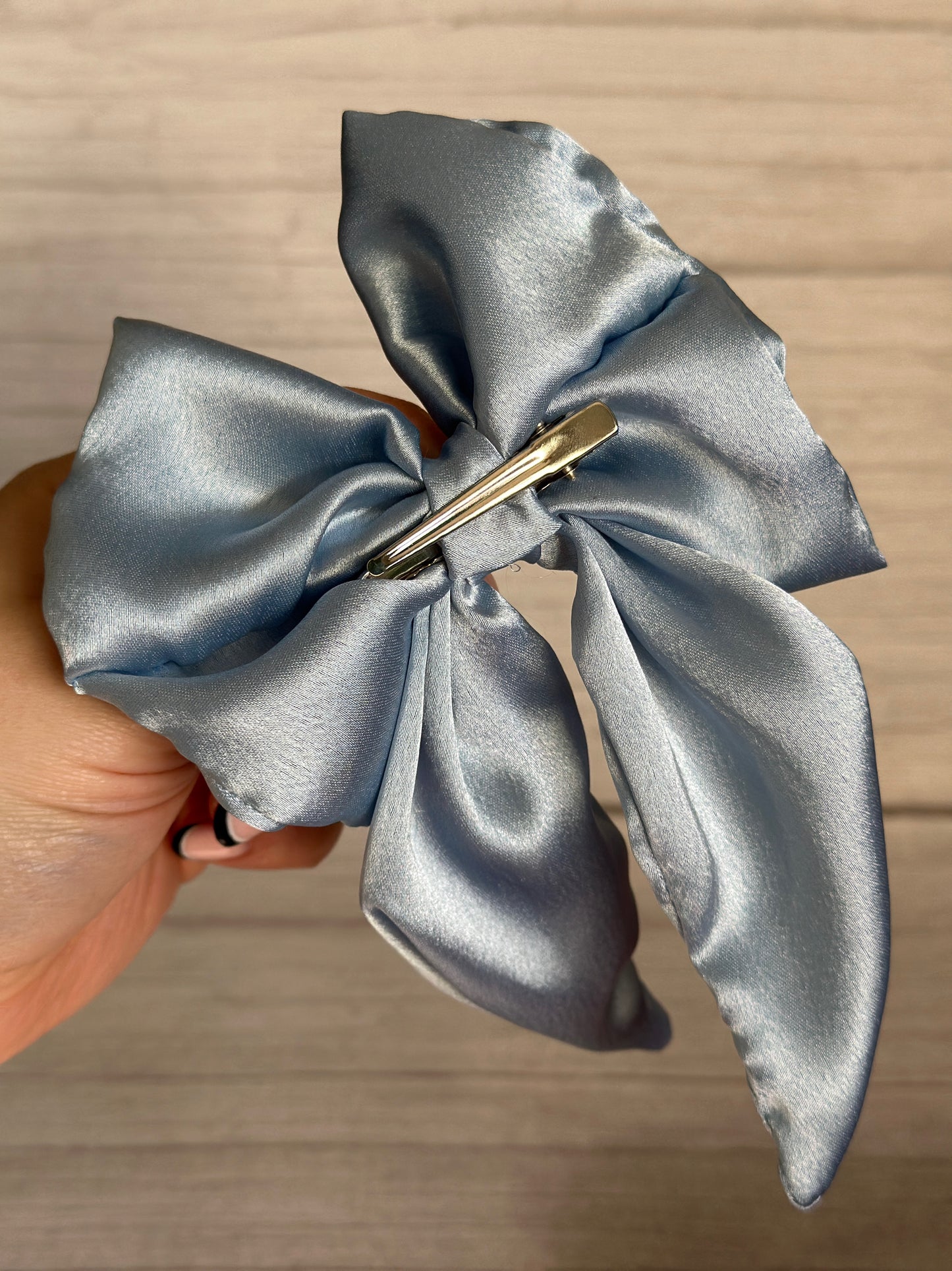 A hand holds the luxurious Silk Sailor Bow Clip in French Blue from Crafts by Kate, showcasing its shiny and textured silk finish. Adorned with a silver hair clip, it offers a handcrafted appearance against a neutral background.