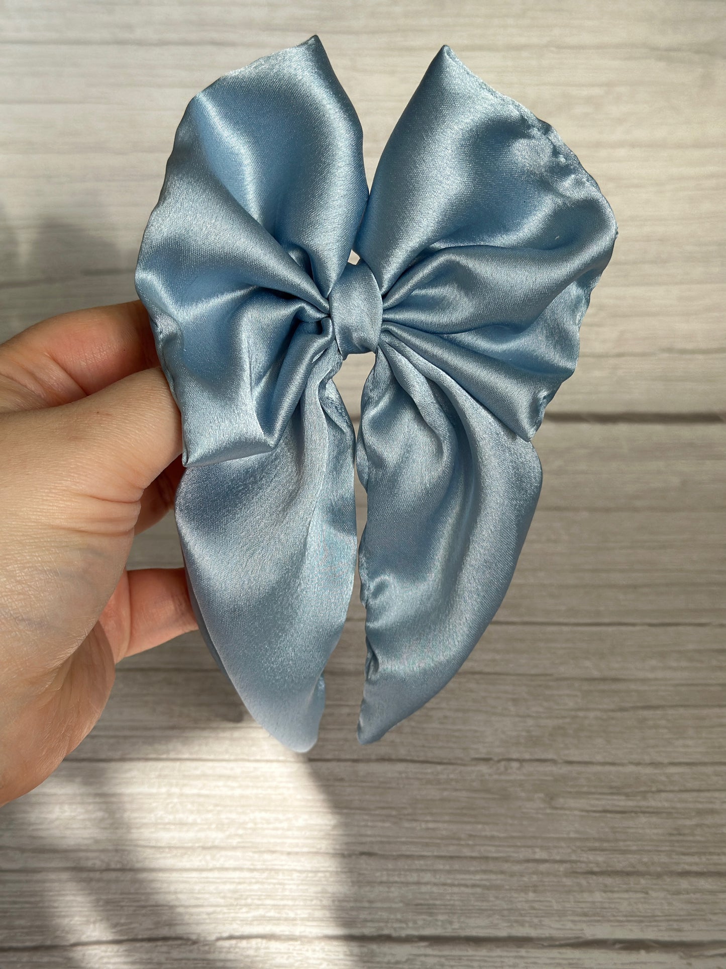 In front of a light wooden background, a hand presents the luxurious Silk Sailor Bow Clip in French Blue by Crafts by Kate. This handcrafted accessory is large and smooth, with long, flowing tails that elegantly enhance any outfit.