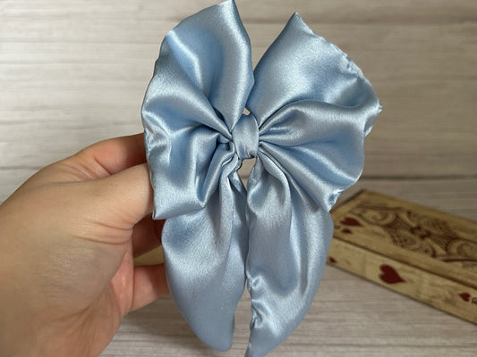 A hand elegantly holds the Silk Sailor Bow Clip in French Blue by Crafts by Kate against a neutral wooden backdrop. In the background, a small handcrafted wooden box decorated with red heart details adds a touch of luxury.