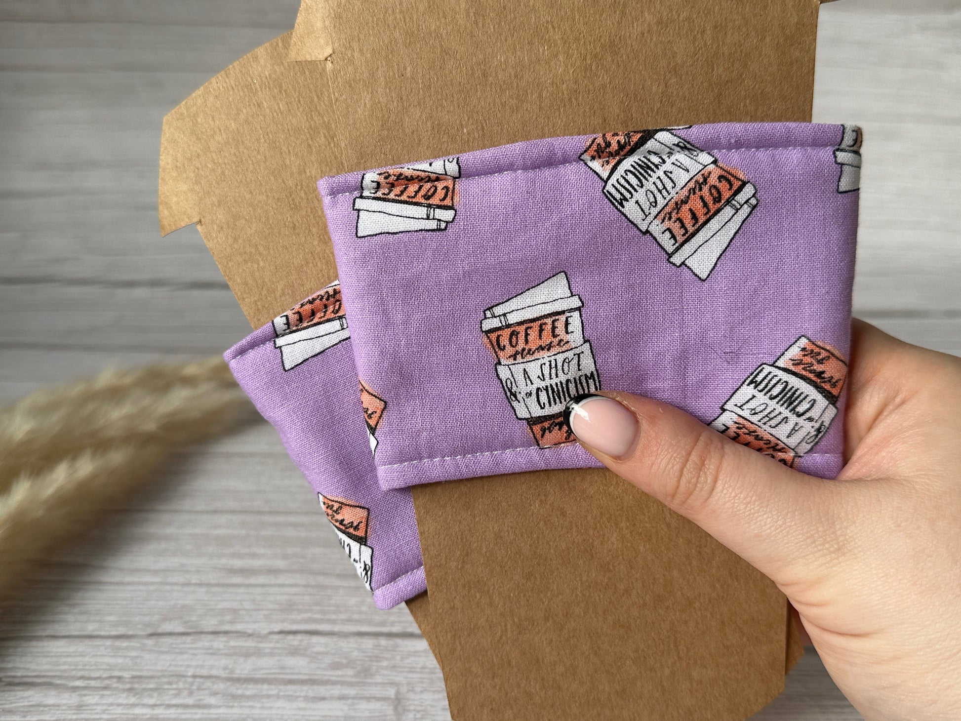 A hand showcasing a Crafts by Kate Cotton Reusable Cup Cozy, designed with a light purple background and adorned with coffee cups and the phrase "Coffee and a Shot of Cynicism." Another identical, eco-friendly cozy is placed on a wooden surface covered with brown paper.