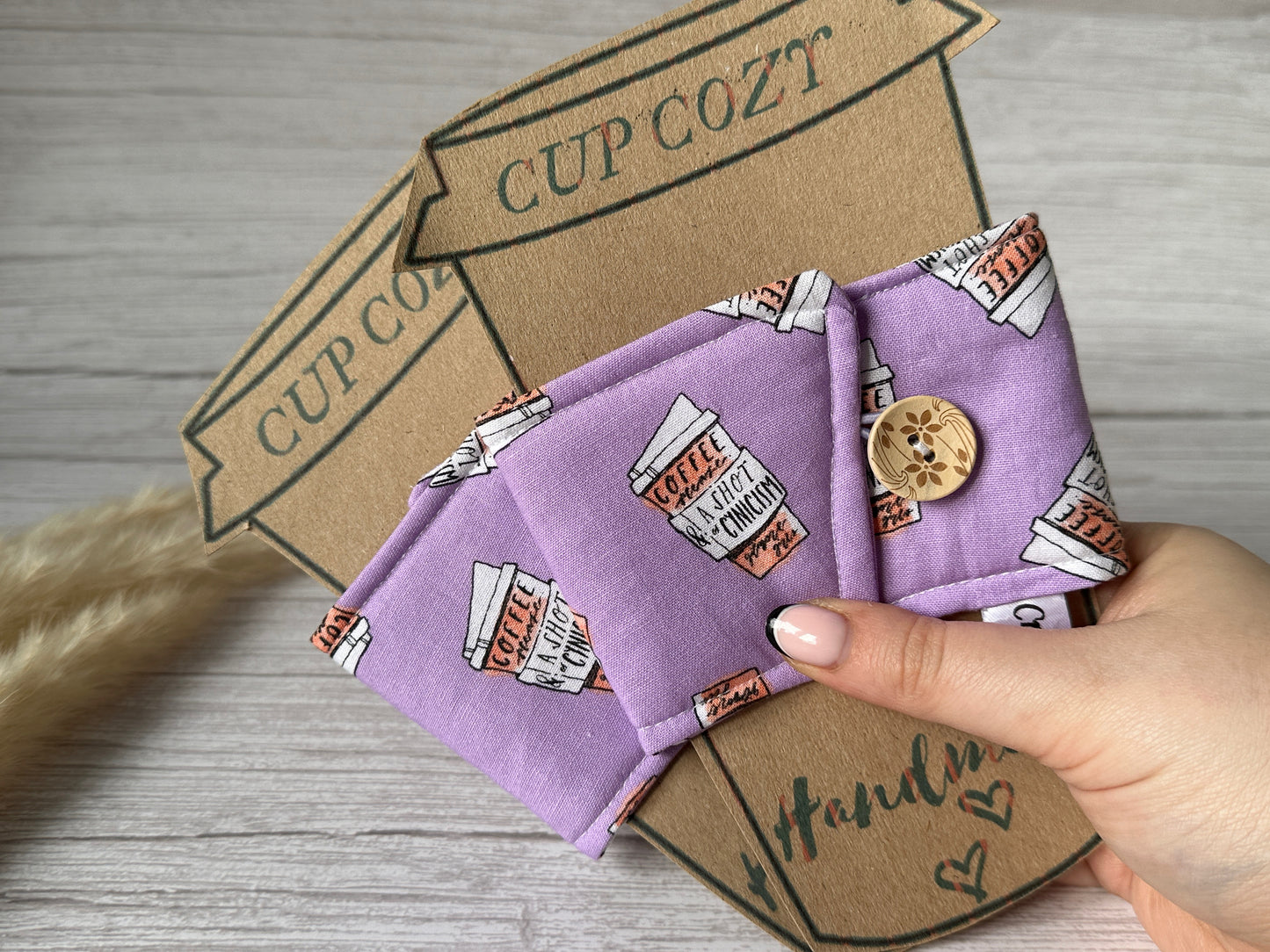 A hand holds a Cotton Reusable Cup Cozy - Gilmore Girls by Crafts by Kate, embellished with a coffee cup pattern and button closure in purple fabric. Behind it stands a brown cardboard cutout shaped like a to-go cup, labeled "Cup Cozy," against a light wooden surface. This handcrafted and environmentally friendly reusable cozy is framed by dried grass on the left side.