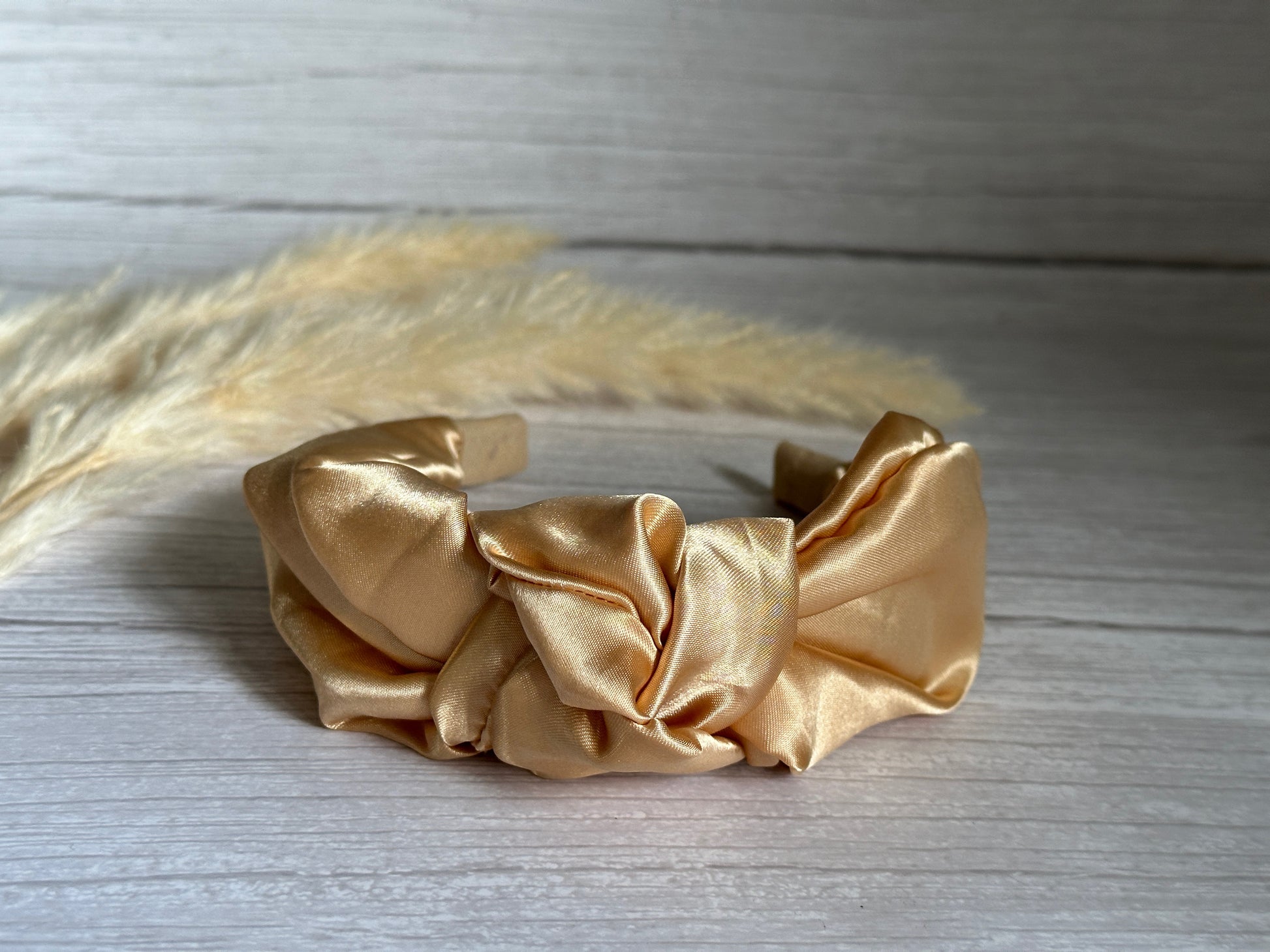 The Silk Knotted Headband - Rich Gold by Crafts by Kate is a luxurious piece featuring an intricate knot design and a gold sheen. It is elegantly displayed on a light wooden surface, with soft, decorative pampas grass providing a delicate background.