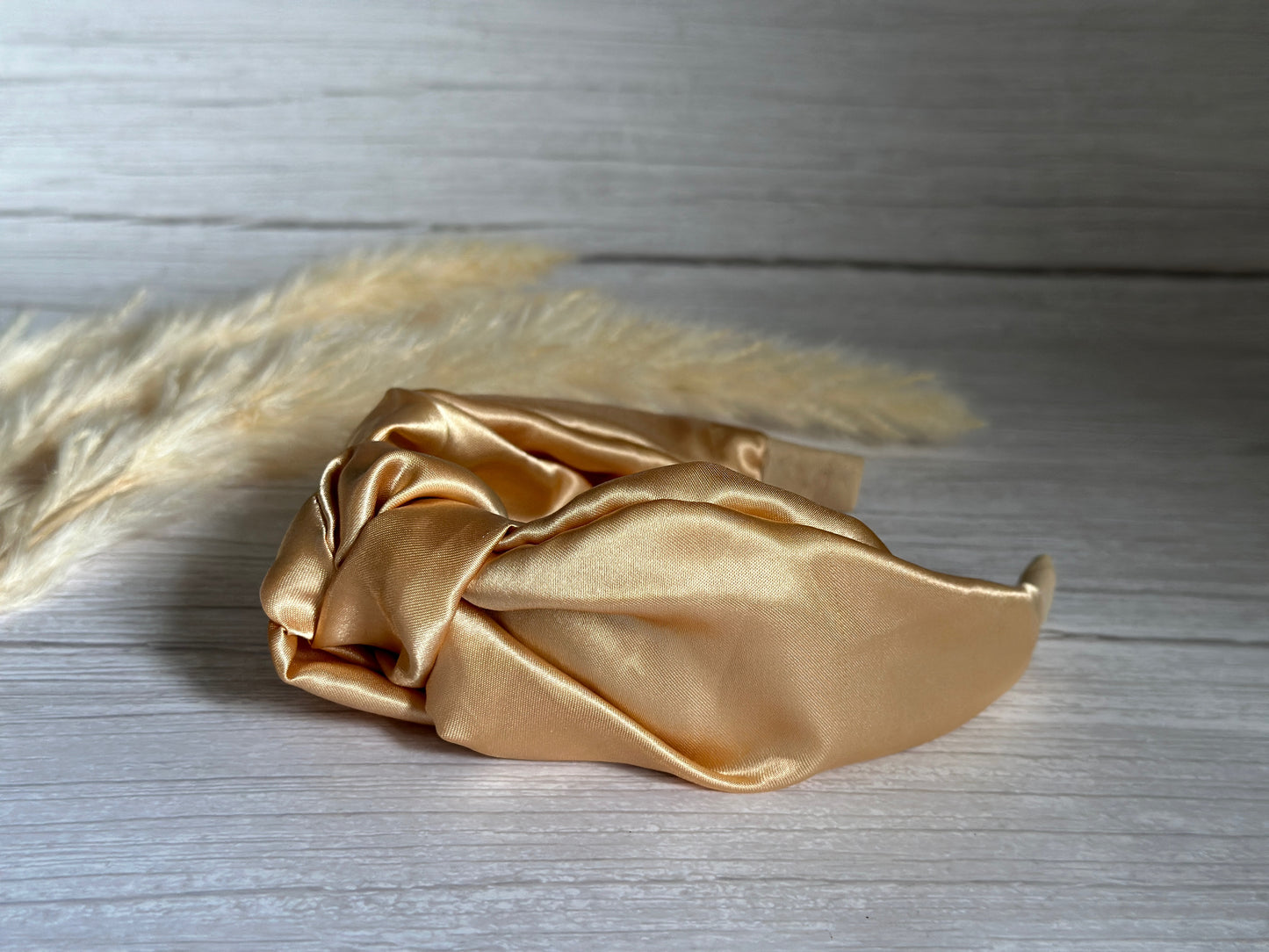 The "Silk Knotted Headband - Rich Gold" by Crafts by Kate rests elegantly on a light wooden surface, accompanied by soft, beige pampas grass, creating a natural and sophisticated composition.