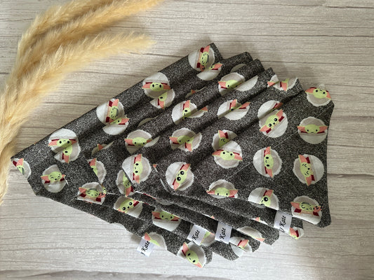 A stack of folded gray cloth napkins made from Crafts by Kate's Cotton Pet Bandana with collar attachment - Star Wars Grogu, featuring small green characters with red hats. The cotton texture is visible on a light wood surface, alongside beige decorative grass stems.