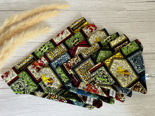 A set of folded fabrics lies on a wooden surface. The Cotton Pet Bandanas with collar attachments from Crafts by Kate, inspired by Harry Potter's Hogwarts house crests, boast intricate designs with dominant colors being green, red, and yellow. Tufts of beige pampas grass are placed to the left of these unique pet accessories.