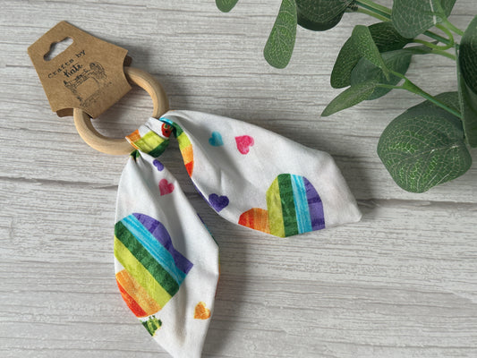 Displayed on a light wood surface with green leaves in the upper right corner, the Crafts by Kate Cotton & Bamboo Baby Teething Rattle - Hungry Caterpillar features a wooden handle and a fabric tie adorned with colorful rainbow heart designs against a white background.