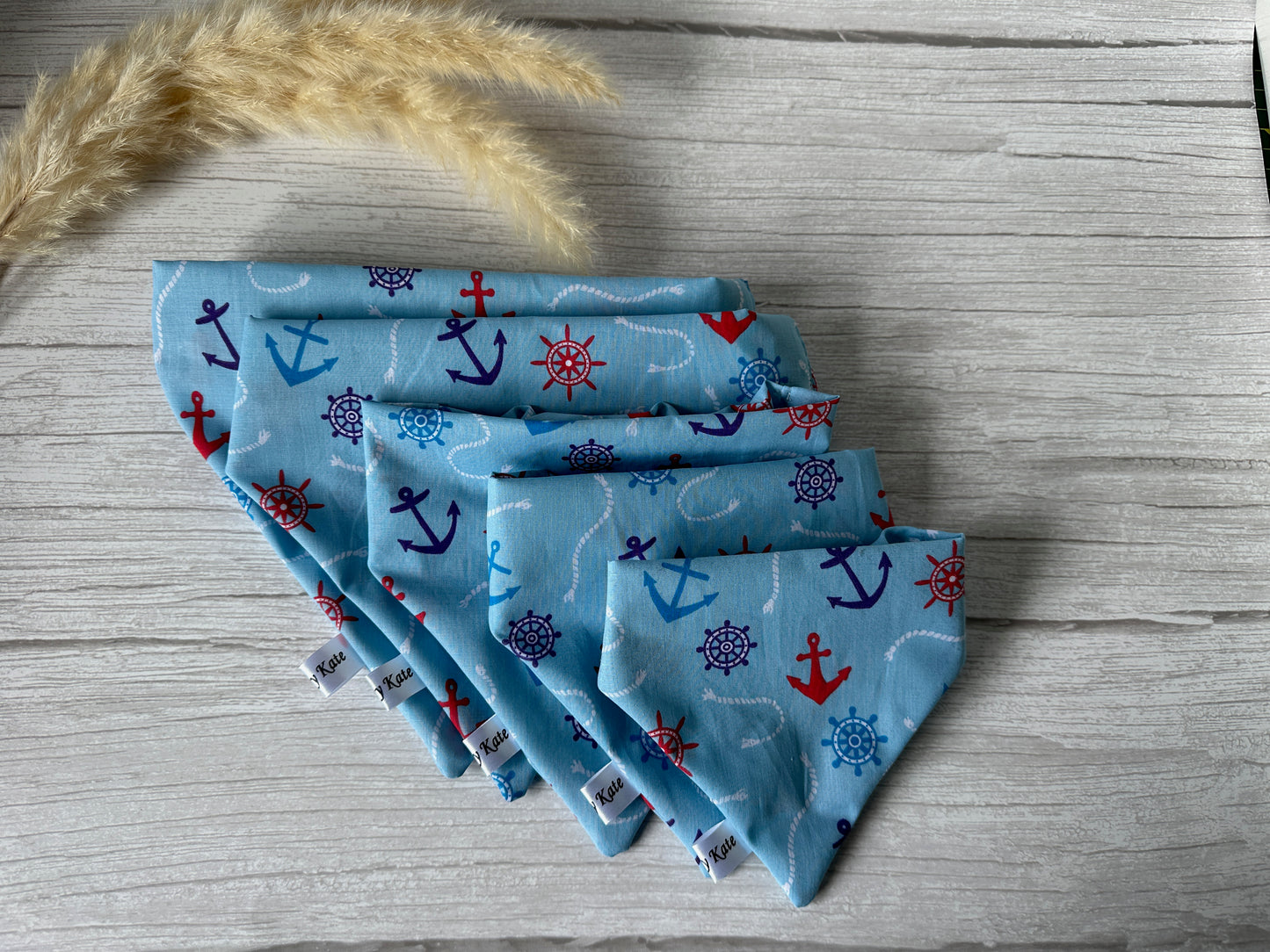 Five Cotton Pet Scrunchie Bandanas - Sailor Print from Crafts by Kate, decorated with nautical patterns including anchors and ship wheels, are beautifully arranged in a fan shape on a wooden surface. Dried pampas grass is positioned above the bandanas. Each handcrafted cotton bandana features a small white tag labeled "Kico.