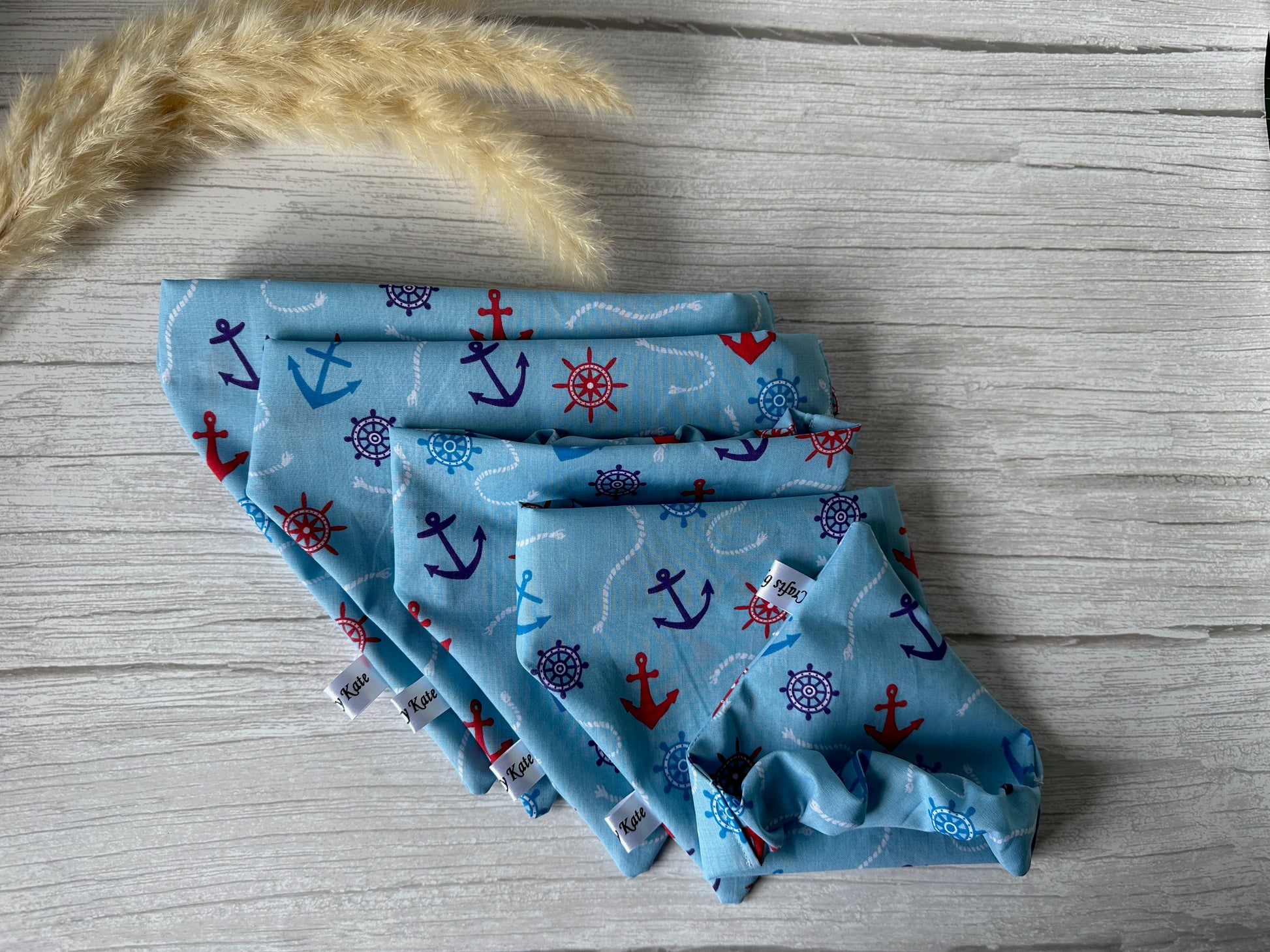 Crafts by Kate's Cotton Pet Scrunchie Bandanas - Sailor Print, featuring colorful nautical designs like anchors and ship wheels, in various sizes, are beautifully arranged on a light wood surface. A sprig of dried grass adorns the top left corner of the image.