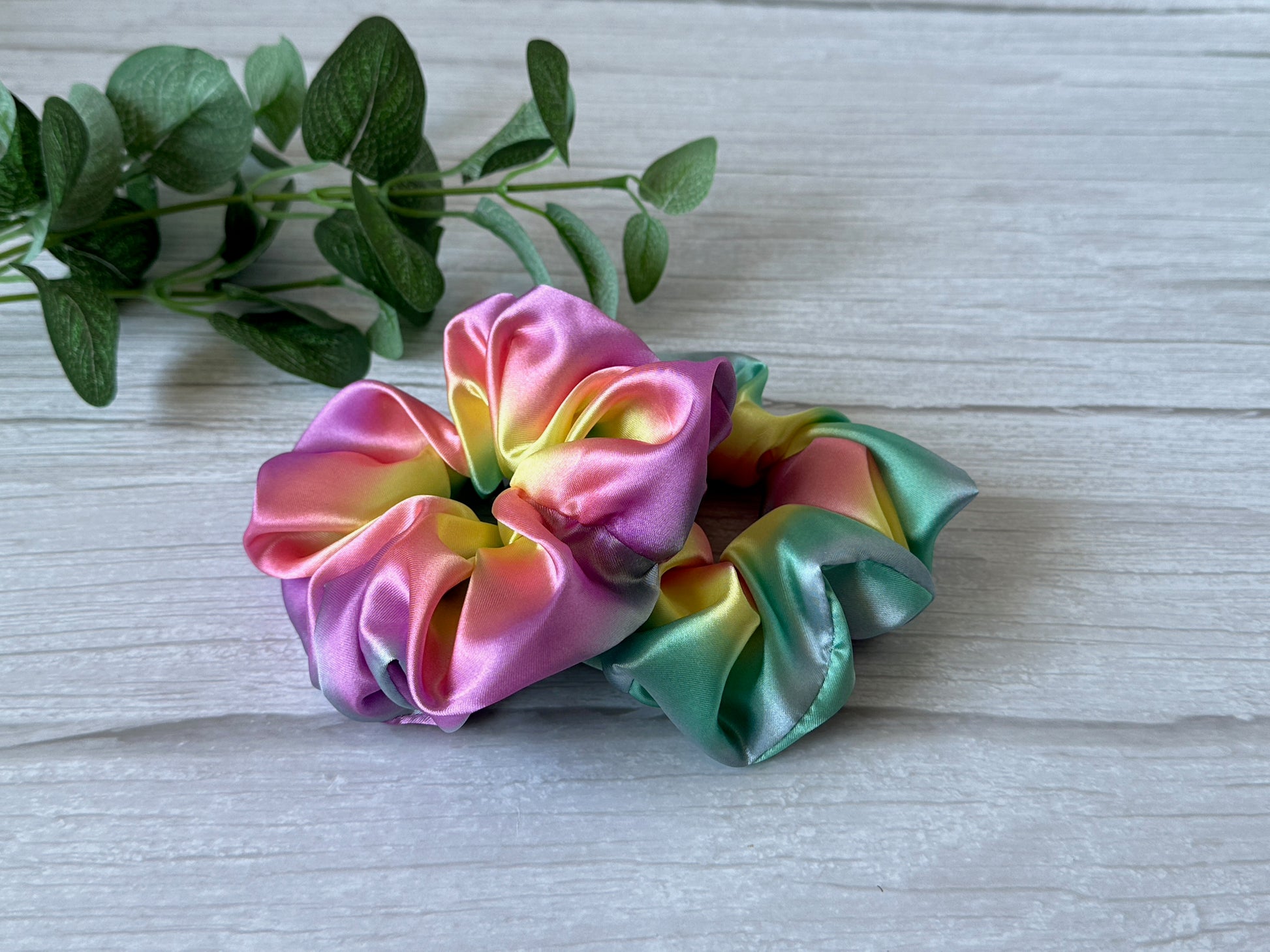 Two vibrant satin scrunchies, featuring the Silk Extra Large Scrunchie in a pastel rainbow of pink, yellow, green, and purple hues from Crafts by Kate, rest on a light wooden surface. A small branch with green leaves lies beside them.