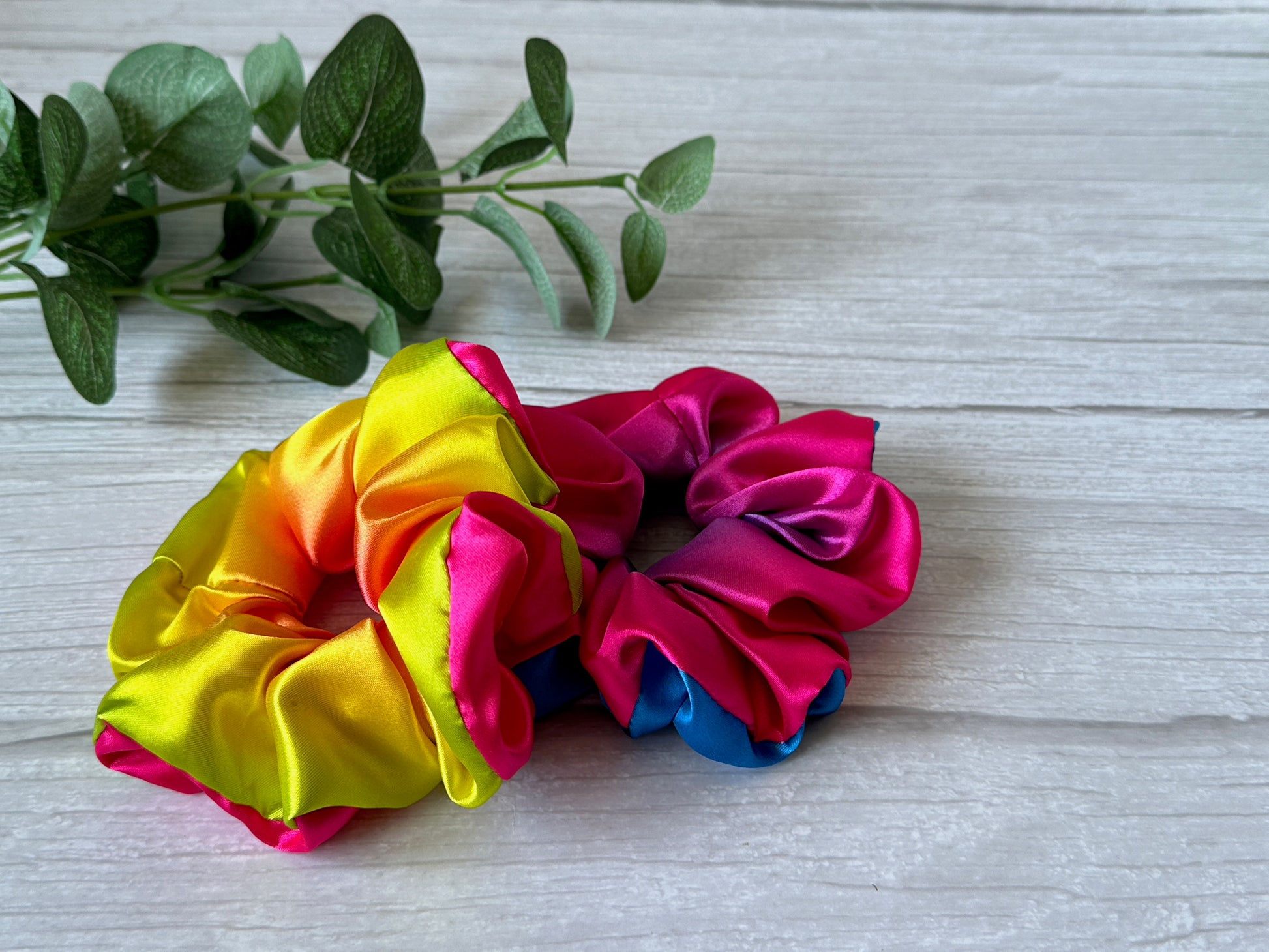 Two vibrant Silk Extra Large Scrunchies by Crafts by Kate, featuring a bright rainbow of colors including yellow, orange, pink, and blue, rest on a light wooden surface. In the background, a sprig of greenery subtly underscores support for the LGBTIQA+ community with their lively tones.