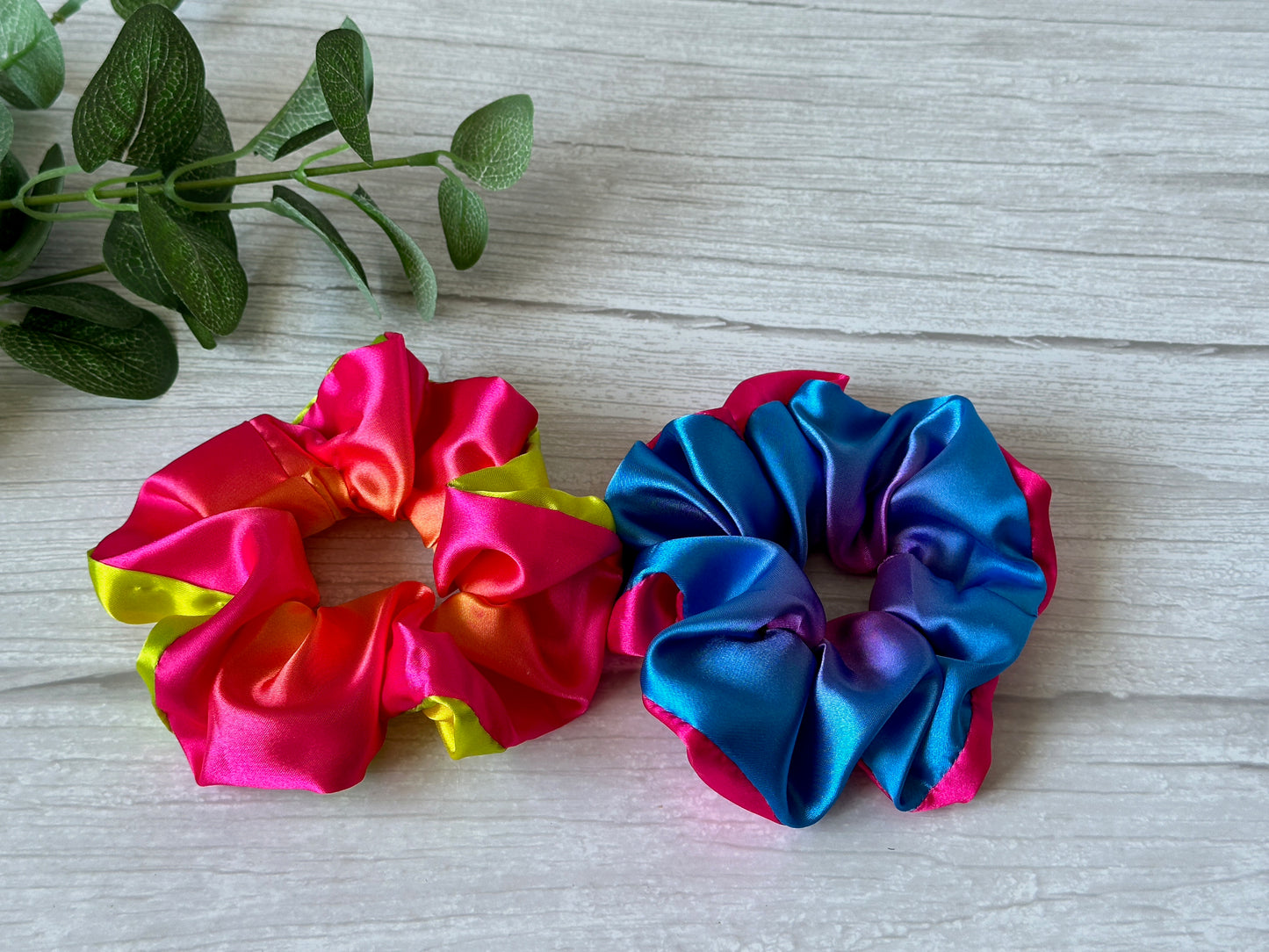 Two Silk Extra Large Scrunchies from Crafts by Kate rest on a white wooden surface. One displays vibrant pink and yellow hues, while the other showcases calming blue and purple tones. A small green plant peeks from the top left corner, infusing the vibrant scene with a touch of nature’s charm. These scrunchies are perfect for supporting personal expression in style every day.