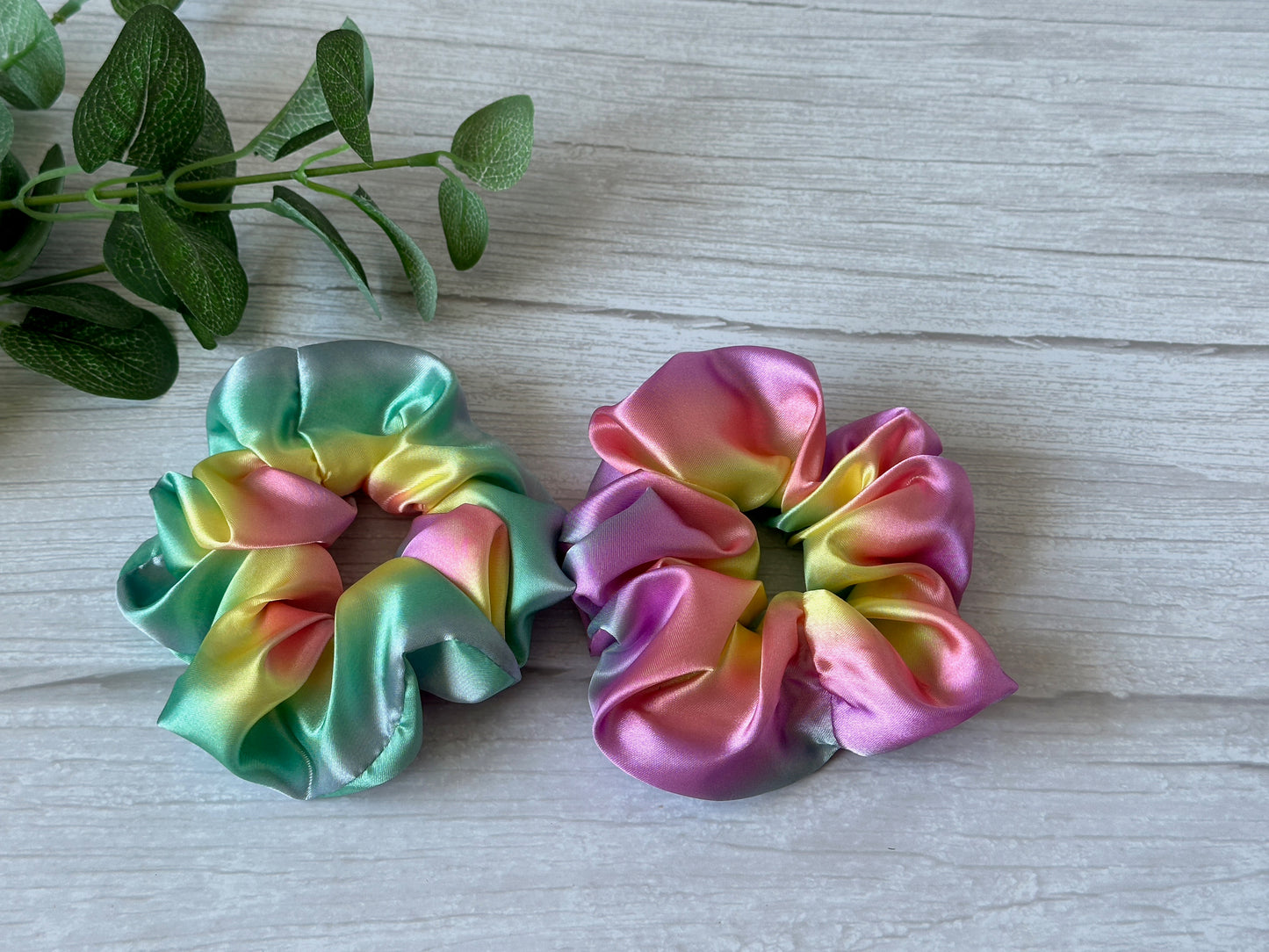 A pair of Silk Extra Large Scrunchies from Crafts by Kate, showcasing a luxurious pastel rainbow gradient in shades of green, pink, yellow, and blue, rest elegantly on a light wood surface. A sprig of green leaves adds a natural touch to the top left corner.