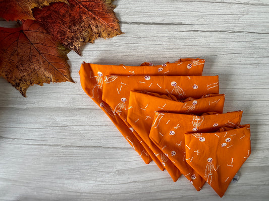 A Cotton Pet Scrunchie Bandana - Dancing Skeletons from Crafts by Kate is neatly folded on a light wooden surface. Autumn leaves arranged in the top left corner enhance its fall theme, while the handcrafted design adds charm, making it ideal for seasonal festivities.