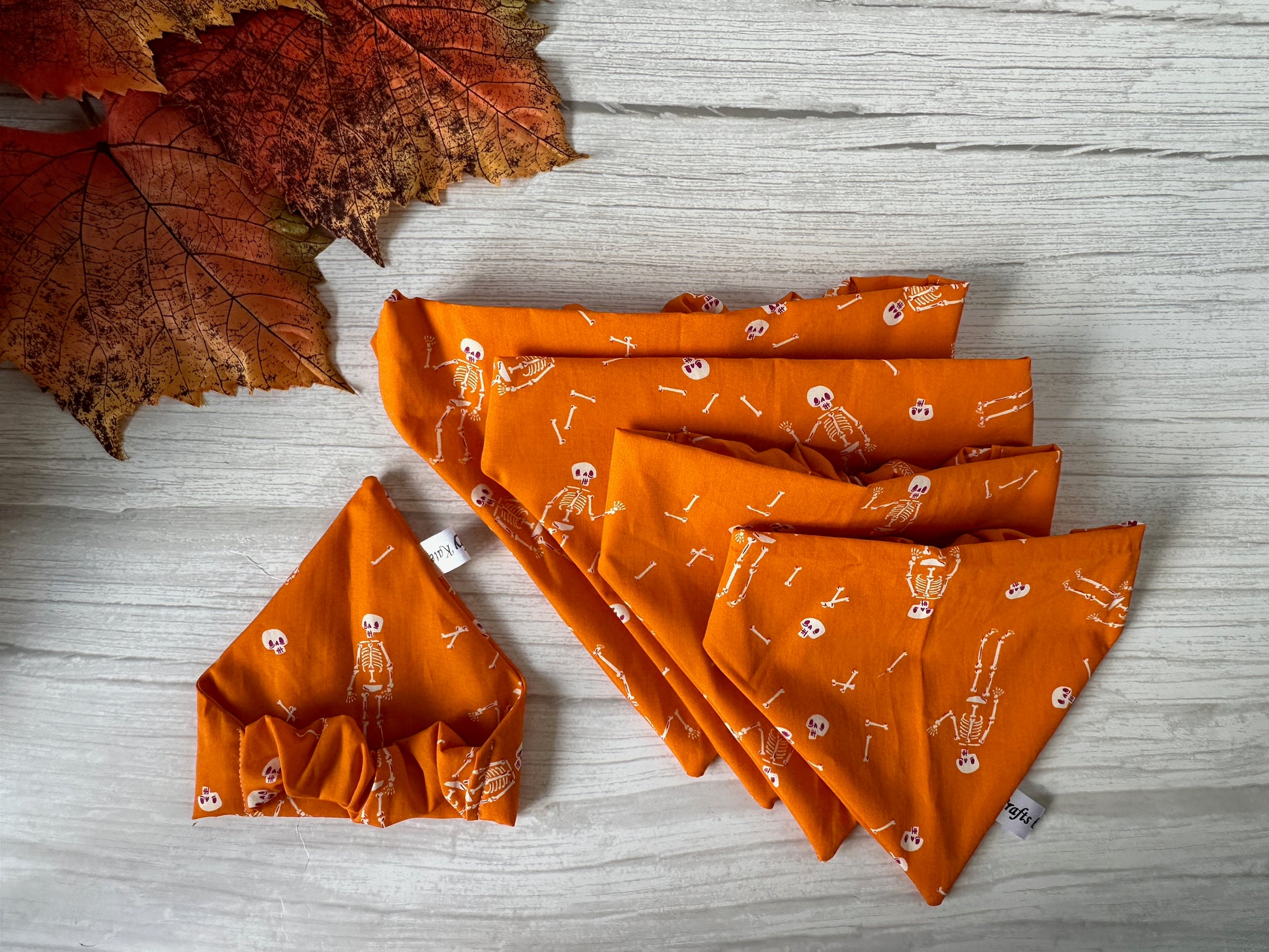 A selection of Crafts by Kate's Cotton Pet Scrunchie Bandanas, featuring dancing skeletons on orange fabric, is neatly arranged on a white wooden surface. A coordinating headband adorned with a bow complements the bandanas beautifully. Handcrafted cotton pet scrunchies add a touch of charm, while autumn leaves are artfully placed in the top left corner.