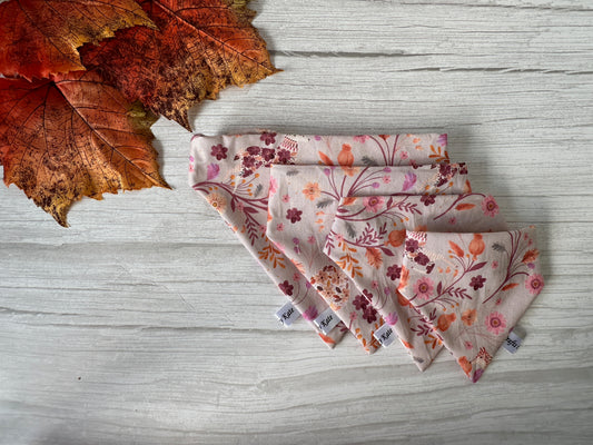 Four neatly folded cloth napkins made from Crafts by Kate's Cotton Pet Scrunchie Bandana in Woodland Floral, featuring shades of pink and orange, are arranged in a fan shape on a light wooden surface. Autumn leaves in hues of orange and red complement the scene to the left, adding a touch of seasonal charm.