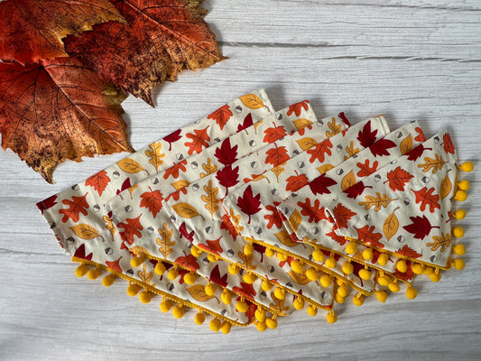 A handcrafted Cotton Pet Bandana with a fall leaf pattern and yellow pom-pom trim, designed by Crafts by Kate, rests on a light wooden surface. Dried orange leaves are artfully arranged in the top left corner. The Autumn Leaves Fabric design highlights red, orange, and yellow foliage.