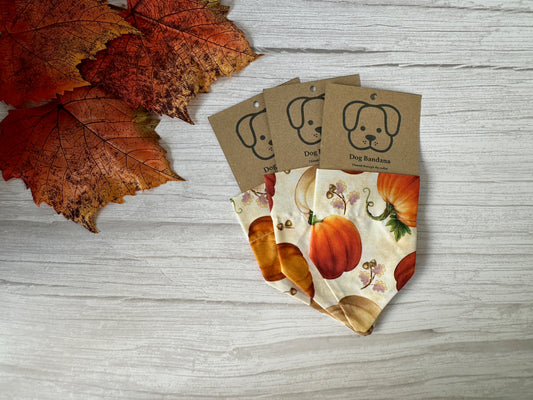 Three Crafts by Kate Cotton Pet Bandanas with collar attachments in Pumpkins fabric are displayed on a white wooden surface, each packaged with a kraft tag featuring a dog illustration, alongside artificial autumn leaves.