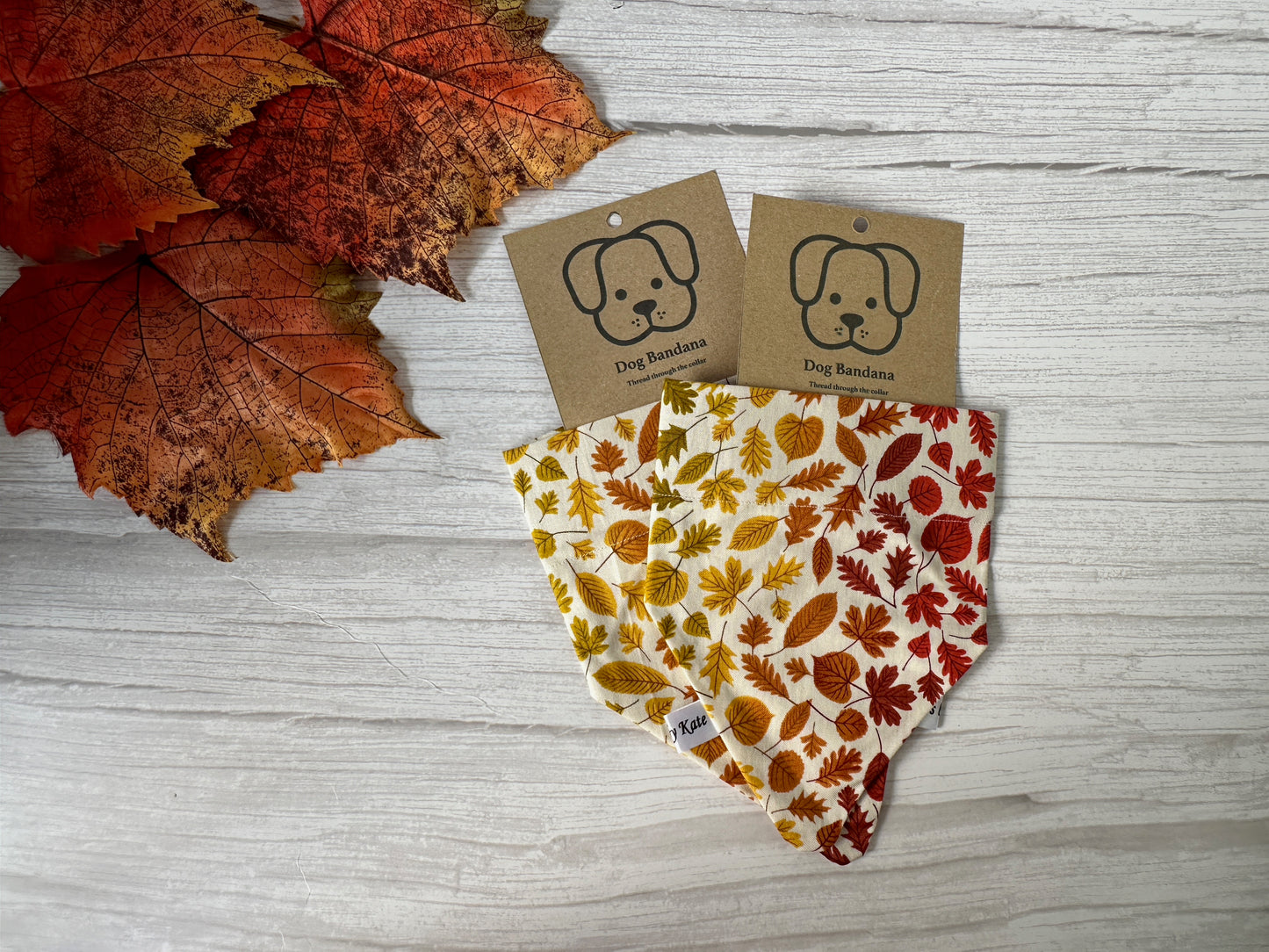 Displayed on a white wooden surface are two Cotton Pet Bandanas with collar attachment from Crafts by Kate, featuring an autumn leaf pattern in vibrant red, orange, and yellow hues. Adjoining them in the top left corner are a few real autumn leaves. Each bandana comes with a tag depicting a dog illustration and is available in various sizes to suit different pets.