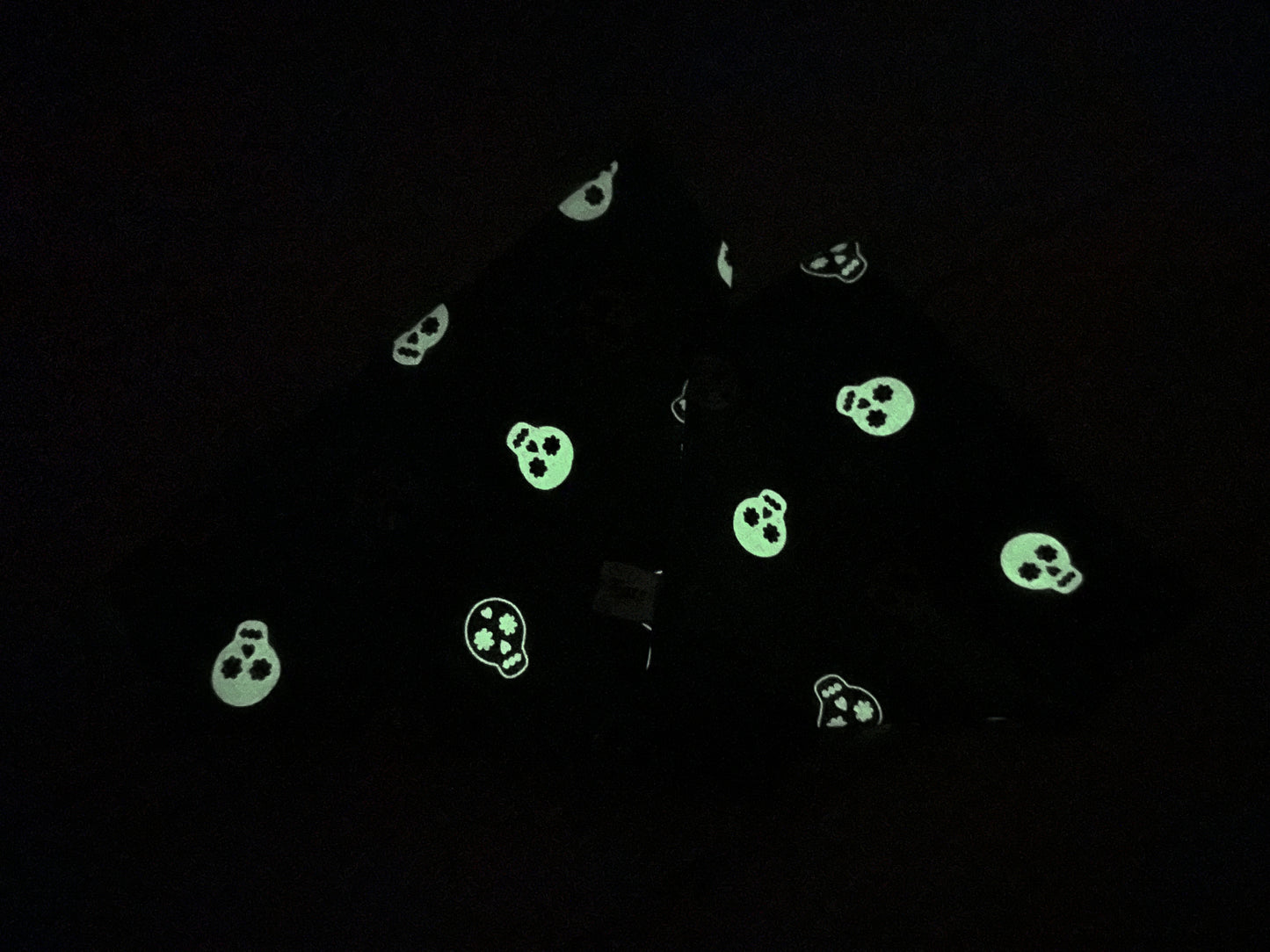 A piece of black fabric from the "Crafts by Kate" line, featuring glow-in-the-dark skeletons with skull patterns scattered across it. The skulls emit a greenish light, illuminating their shapes against the dark background. Perfect for creating the handcrafted "Cotton Pet Scrunchie Bandana - Glow in the Dark Skeletons." The fabric appears to be folded or draped.