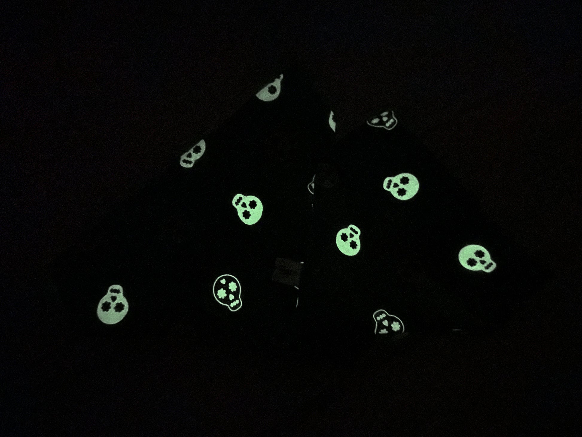 A piece of black fabric from the "Crafts by Kate" line, featuring glow-in-the-dark skeletons with skull patterns scattered across it. The skulls emit a greenish light, illuminating their shapes against the dark background. Perfect for creating the handcrafted "Cotton Pet Scrunchie Bandana - Glow in the Dark Skeletons." The fabric appears to be folded or draped.