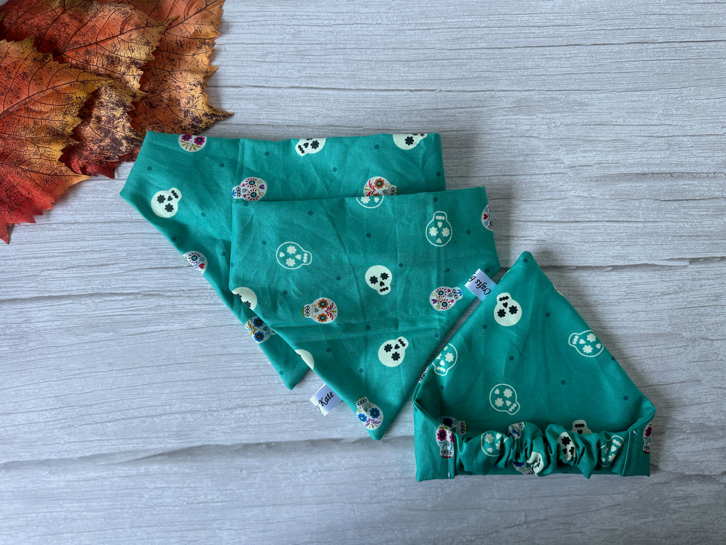 Three teal-colored cloth face masks featuring vibrant sugar skull designs are arranged on a wooden surface, showcasing both their front and back sides. A few autumn leaves in the upper left corner bring a touch of fall charm, reminiscent of the handcrafted "Cotton Pet Scrunchie Bandana - Glow in the Dark Skeletons" by Crafts by Kate.