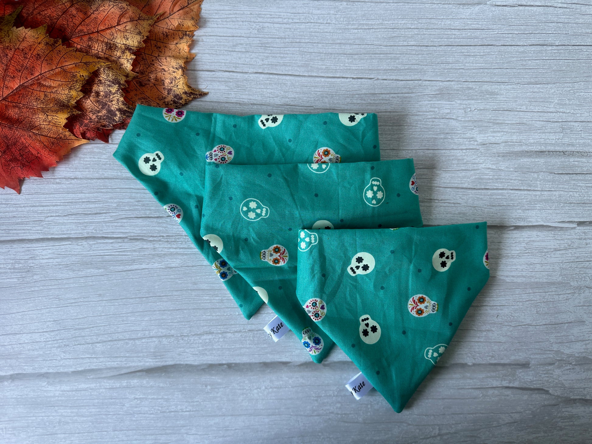 Three Cotton Pet Scrunchie Bandanas - Glow in the Dark Skeletons by Crafts by Kate, adorned with various colorful skull patterns, are neatly arranged on a gray wooden surface. The top left corner features several autumn leaves in shades of red and orange, adding a seasonal touch to the scene.