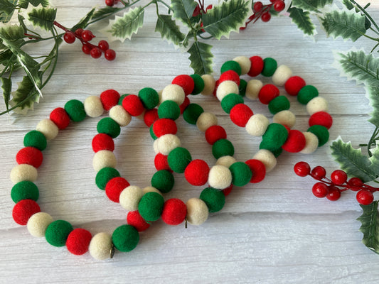 Four PomPom Pet Collars by Crafts by Kate, made of red, green, and white felt balls, sit on a wooden surface. Surrounded by holly leaves and berries, these handcrafted collars offer a festive touch as charming pet accessories for the holiday season.