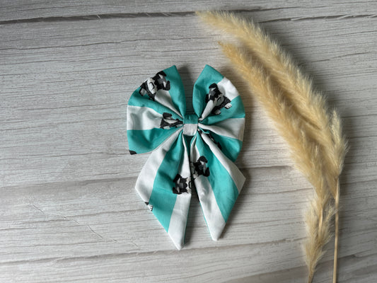 The Cotton Sailor Bow Clip - Striped Schnauzers by Crafts by Kate features luxury turquoise and white stripes with small cow prints. Adorned on a wooden surface, three dried pampas grass stalks to the right enhance its exquisite appeal.