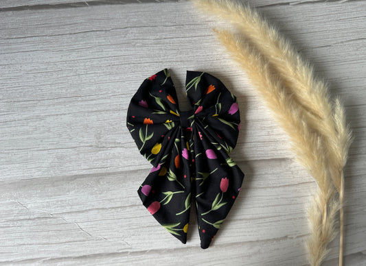 The Cotton Sailor Bow Clip - Tulips by Crafts by Kate is a luxury bow with multicolored floral patterns reminiscent of handcrafted tulips, resting on light wood. Wispy beige pampas grass stalks elegantly complement its design.