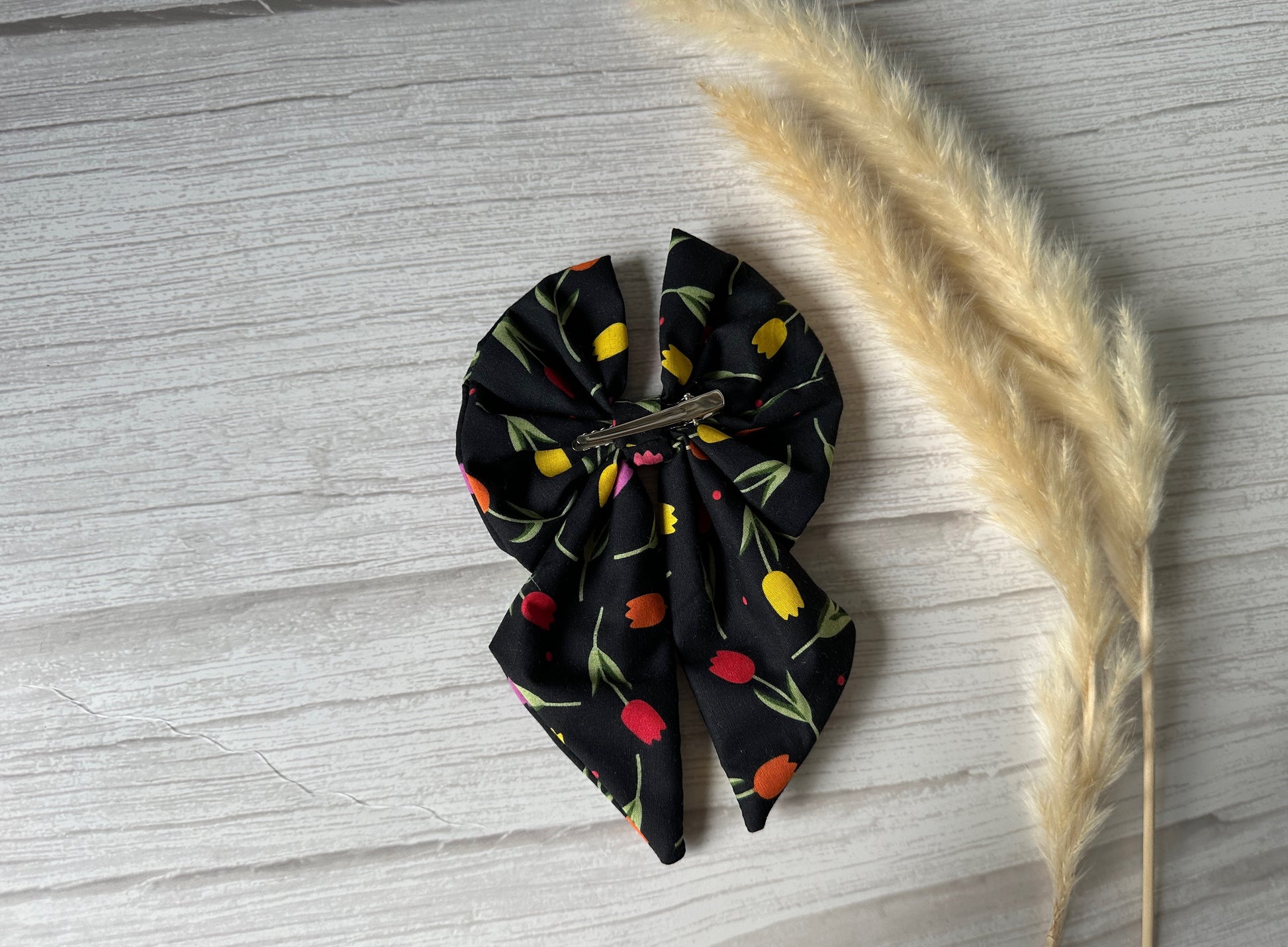 The Cotton Sailor Bow Clip - Tulips by Crafts by Kate, with its colorful floral pattern of red, yellow, and orange tulips, rests on a wooden surface beside two beige fluffy pampas grass stems.