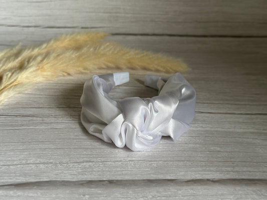 A Silk Knotted Headband in Ice White by Crafts by Kate rests on a light wooden surface, with fluffy beige pampas grass in the background, evoking elegance and style.