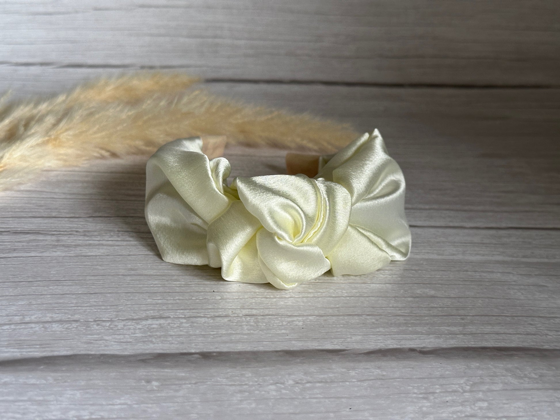 A luxurious Ivory Silk Knotted Headband by Crafts by Kate is elegantly placed on a wooden surface, with beige pampas grass blurred in the background. This handcrafted accessory boasts a shiny texture, flawlessly arranged at the center.