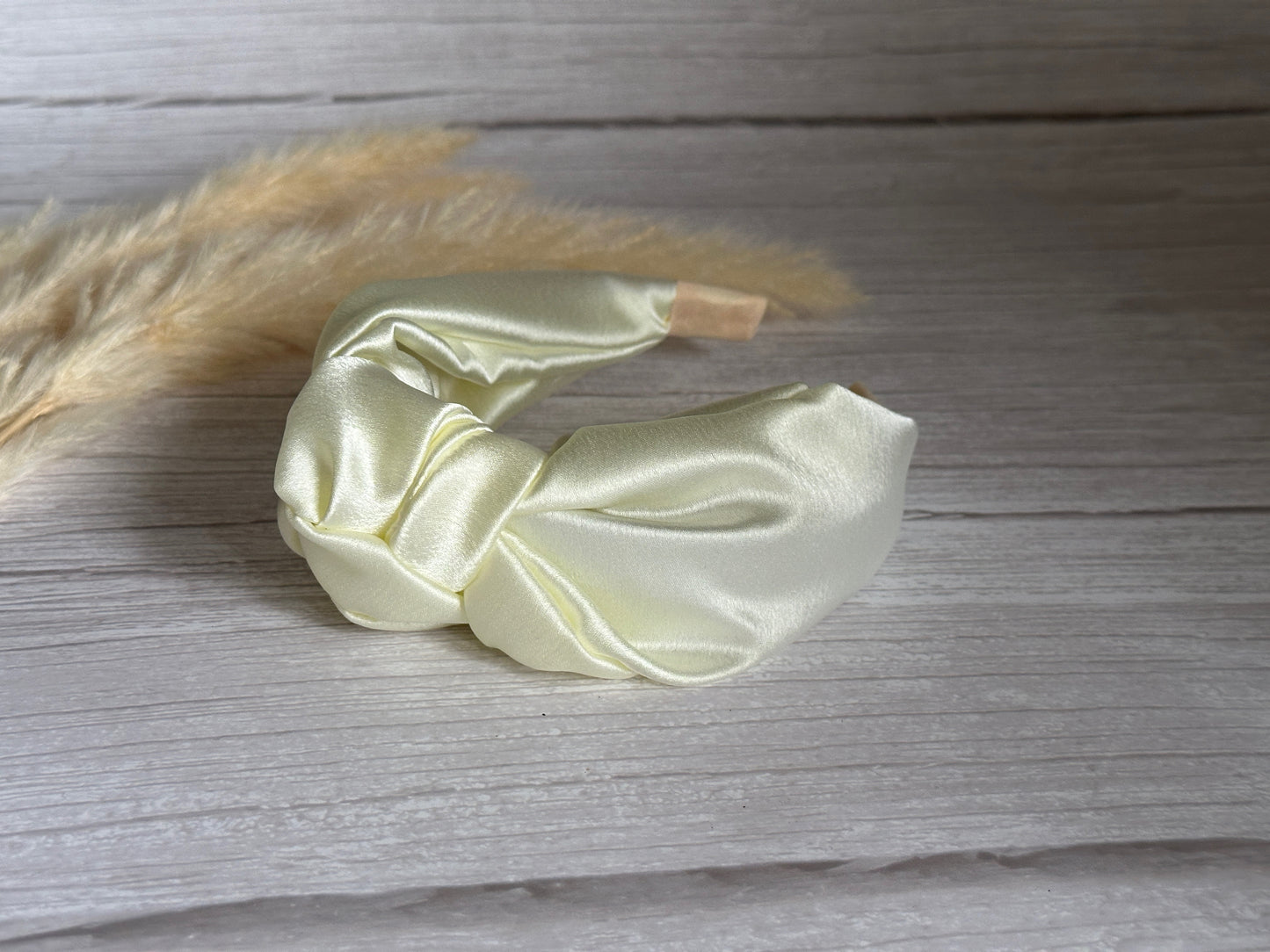 The Silk Knotted Headband - Ivory by Crafts by Kate is elegantly twisted and placed on a light wooden surface, with dried pampas grass in the background.